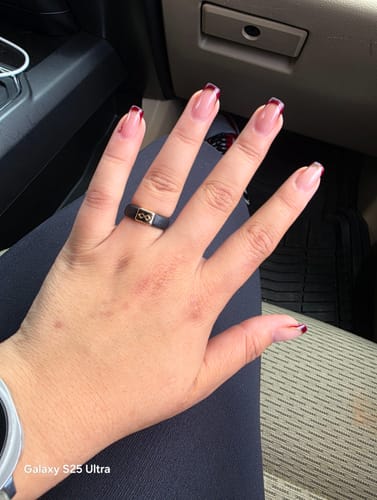 A reviewer's hand wearing the black silicone wedding ring with a gold plate featuring an infinity symbol, seen in a car.