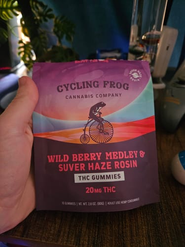 A reviewer holds the purple pouch of Wild Berry Medley Rosin Gummies in their hand, with a bong in the background.