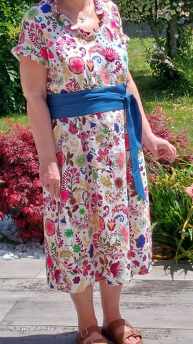 Cliente portant la robe Acacia à imprimé floral avec une large ceinture bleue, vue de trois-quarts dans un jardin.