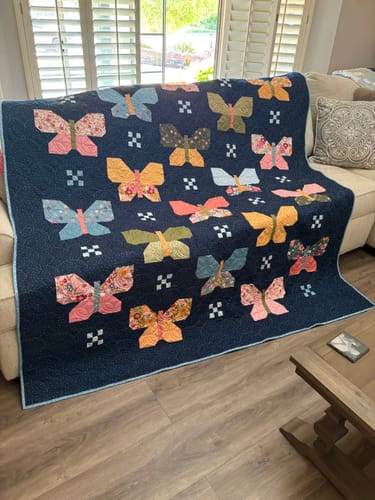 Reviewer's finished Flutter quilt with colorful patchwork butterflies on a dark blue background, draped over a sofa.