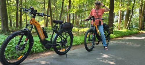 Customer's two orange Lankeleisi MG600 Lite e-bikes on a paved path in a sunny, wooded area.