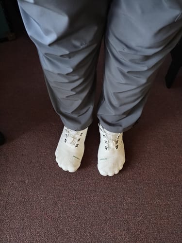 Customer standing indoors wearing white Free I Barefoot Shoes on a dark carpet.