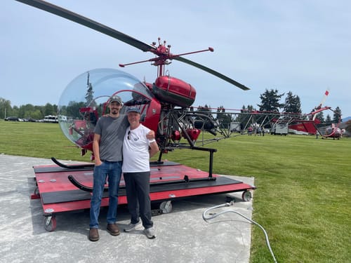 Customer and another person standing in front of a small red helicopter on a grassy field.