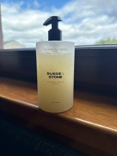 Reviewer's Suede & Stone Body Wash in a frosted dispenser bottle, sitting on a wooden windowsill.