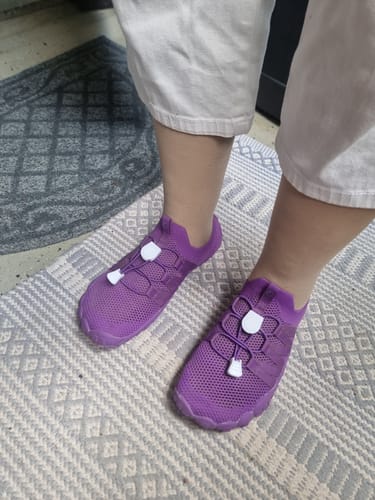 Asiakas esittelee violetteja Vapauta Varpaat™ Paljasjalkakenkiä jaloissaan kuviollisen maton päällä.
