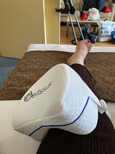 Cliente utilisant le coussin orthopédique pour genoux Cellsius entre ses jambes, avec des béquilles à l'arrière-plan.