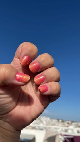 Reviewer's hand showing Sunset Breeze nail wraps with a coral and peachy gradient against a bright blue sky.