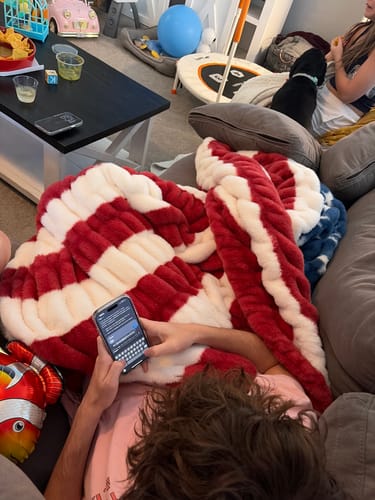 A person lying on a couch covered in the plush, red and white striped Hugs Flag Blanket while using a phone.