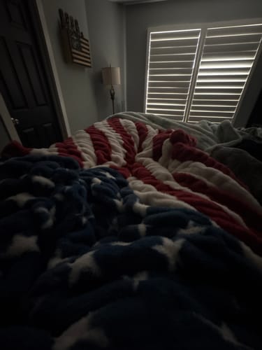 Reviewer's plush Hugs Flag Blanket draped over a bed in a dimly lit room, showing its thick texture.