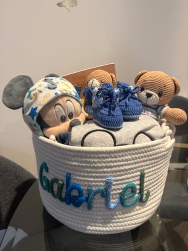 Customer's white personalized knitted basket with the name 'Gabriel' in multicolor yarn, filled with baby gifts and toys.