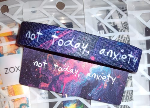 Customer's Not Today, Anxiety Bracelet laid over its matching card, both featuring a colorful galaxy design.