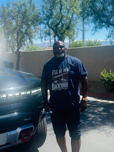 Customer standing outdoors wearing the dark blue 'I’ll Be In The Backyard' shirt.
