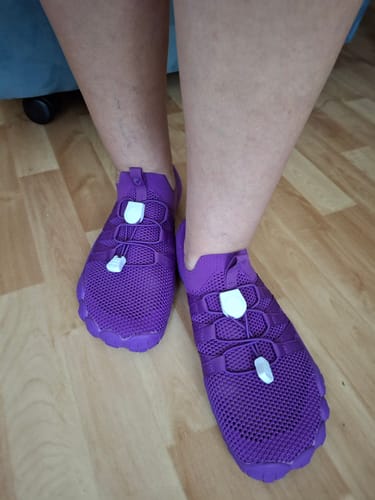 Asiakkaan jalassa violetit Vapauta Varpaat™ Paljasjalkakengät -kengät puisella lattialla.