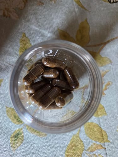 Reviewer's clear jar filled with capsules of ground CBD Small Buds, seen from above on a patterned surface.