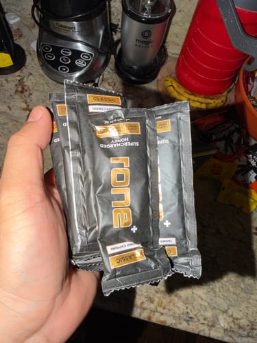 Customer's hand holding several packets of Classic SuperCharged Honey over a kitchen counter with blenders in the background.