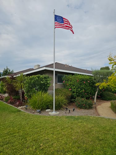Customer's Betsy Ross Flag flying on a tall flagpole in the front yard of a house with landscaped bushes.