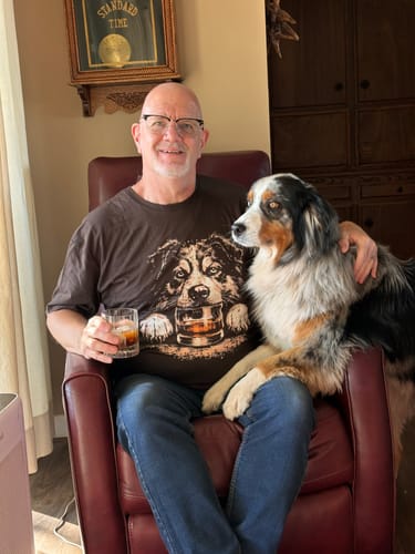 Customer wearing the Whiskey Australian Shepherd T-Shirt while sitting with his dog and holding a glass of whiskey.