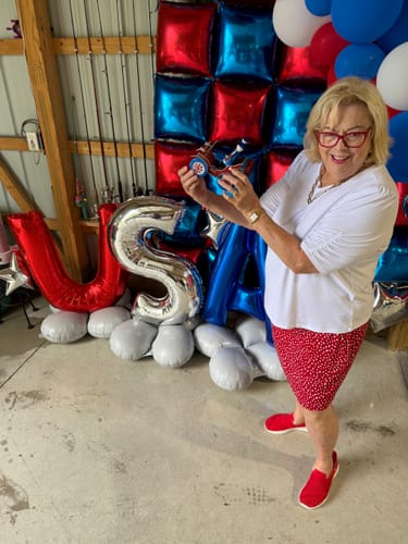 Reviewer smiling while holding a small object in front of patriotic USA-themed balloons.