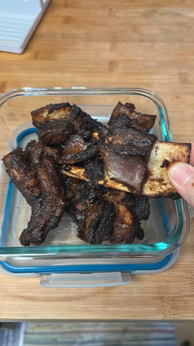 Customer holding a piece of roasted Halal Veal Pulao Meat on the bone over a glass container full of cooked meat.