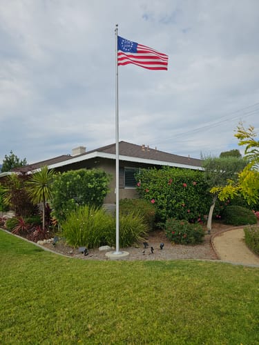 Customer's Betsy Ross Flag flying on a tall flagpole in the front yard of a house with landscaped bushes.