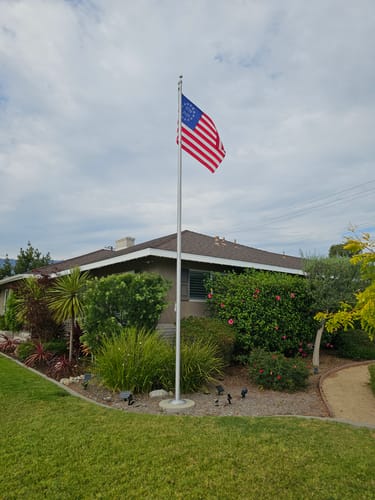 Customer's Betsy Ross Flag flying on a tall flagpole in the front yard of a house with landscaped bushes.