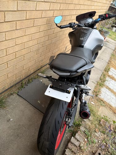 Reviewer's Universal Adjustable Flip Bracket installed on the rear of a grey and black motorcycle, parked next to a brick wall.