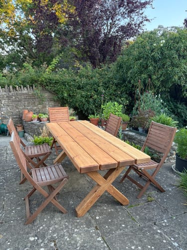 Customer's sturdy X Frame Garden Table on a patio, surrounded by six wooden chairs and lush greenery.