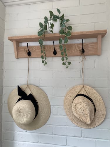 Customer's elegant oak coat rack with shelf mounted on a white brick wall, holding two hats and a plant.
