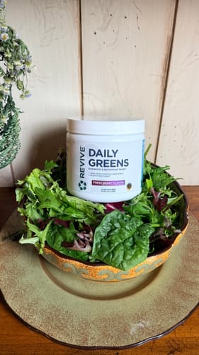 Customer's white tub of Revive Daily Greens sitting in a large bowl of fresh salad greens.