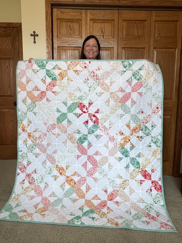 A customer proudly holds up her finished Bloomcross Quilt, showing the colorful floral cross-and-bloom pattern.