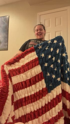 Customer smiling while holding up the large, plush Hugs Flag Blanket indoors.