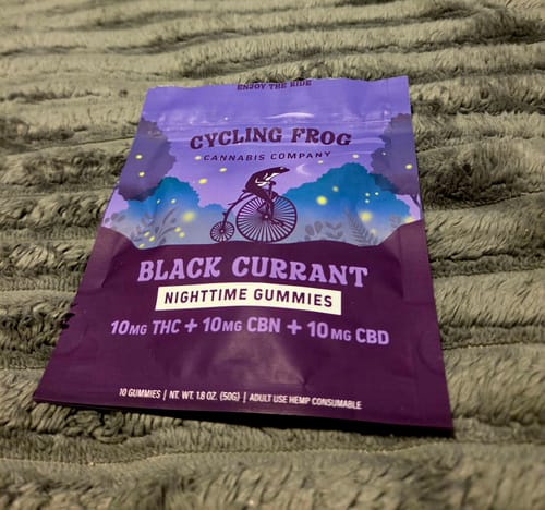 A customer's purple pouch of Cycling Frog Black Currant Nighttime Gummies resting on a gray textured blanket.