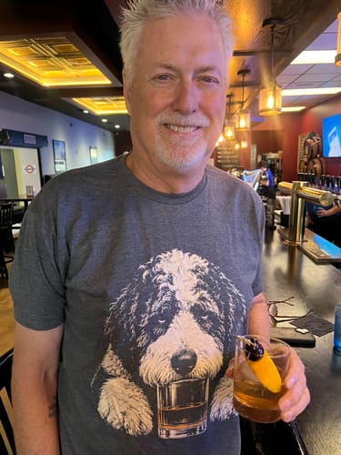 Customer wearing the grey Whiskey Dog Sheepadoodle T-Shirt while at a bar holding a cocktail.