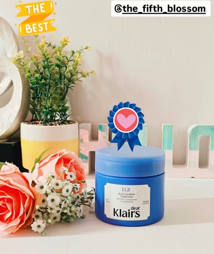 Reviewer's blue tub of EGF Blue Calming Toner Pad displayed on a white surface next to flowers and a plant.