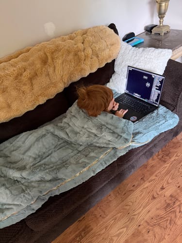 Customer's child snuggled in the cozy Plush Mist Sleeping Bag while using a laptop on a couch.