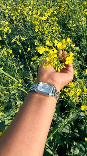 Customer's arm wearing a silver, square-faced watch while holding yellow flowers in a field.