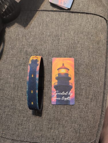 Customer's Guided By Your Light Bracelet and its matching collector's card, both with a lighthouse design, on a gray sofa.