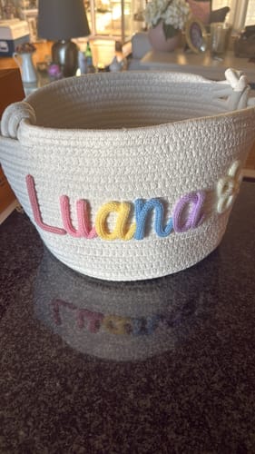 Customer's personalized white knitted basket with the name Luana in pastel yarn, sitting on a dark surface.