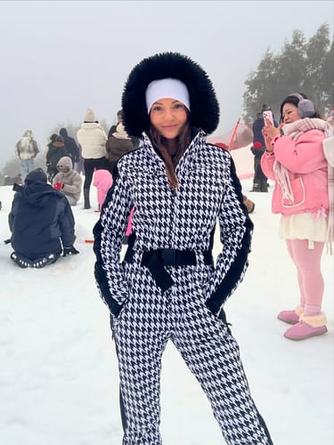 Reviewer wearing a black and white houndstooth one-piece ski suit with a fur-trimmed hood in a snowy outdoor setting.