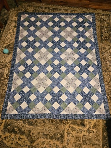Customer's completed Belvoir Blue Quilt Kit with a lattice design in blue, gray, and white on a patterned rug.
