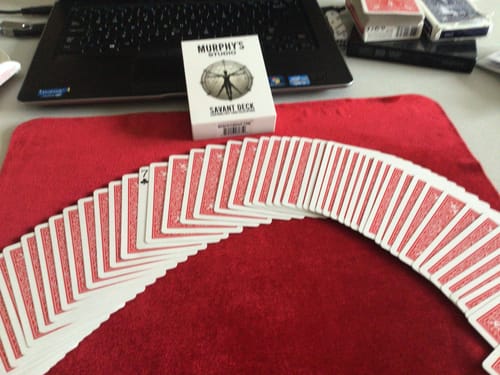 Reviewer's Savant Deck box sitting on a desk next to a fanned-out red-backed card deck on a red velvet mat.