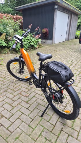 Reviewer's orange and black Lankeleisi MG600 Lite e-bike parked on a brick driveway, shown from a side-rear angle with a bag on the rack.
