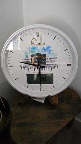 Customer's white Sajada Azan Clock with a Kaaba illustration, resting on its wooden stand.