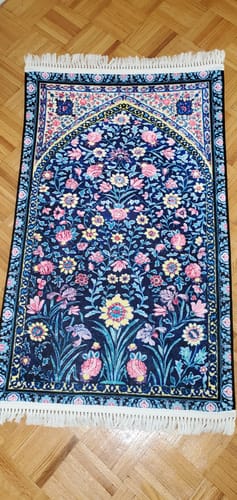 Customer's Noor al-Qalb Padded Prayer Mat in dark blue with a vibrant floral pattern and white fringe, laid on a wooden floor.