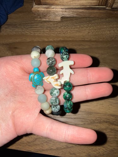 Customer's hand holding three colorful beaded Shark Week bracelets with shark charms.