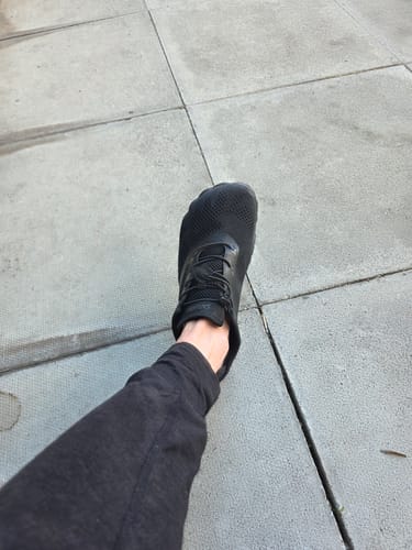 A customer's top-down view of their foot in a black Smart I barefoot shoe on a paved walkway.