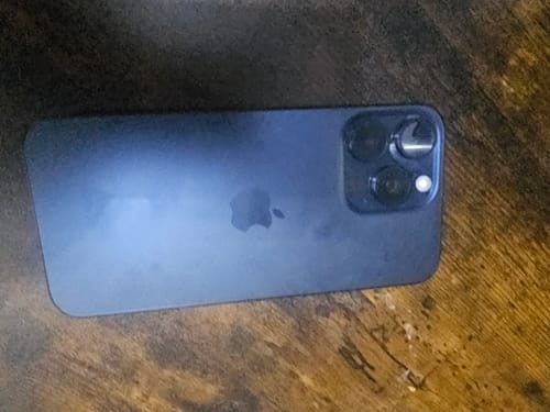 Customer's dark-colored Apple iPhone 15 Pro Max showing the back and camera system on a wooden surface.