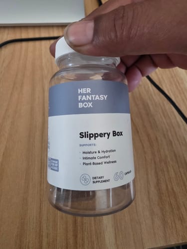 Customer's hand holding a clear bottle of Slippery Box dietary supplement against a wooden surface.