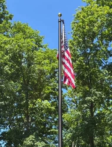 Close-up of a customer's dark commercial flagpole with an American flag set against green trees.