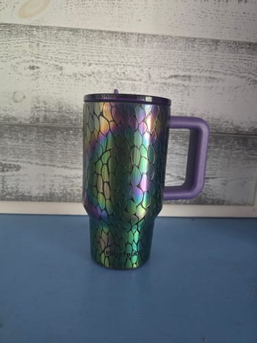 Reviewer's Thicc Buddy 16oz mug with iridescent mermaid scale pattern, purple lid, and handle on a blue surface.